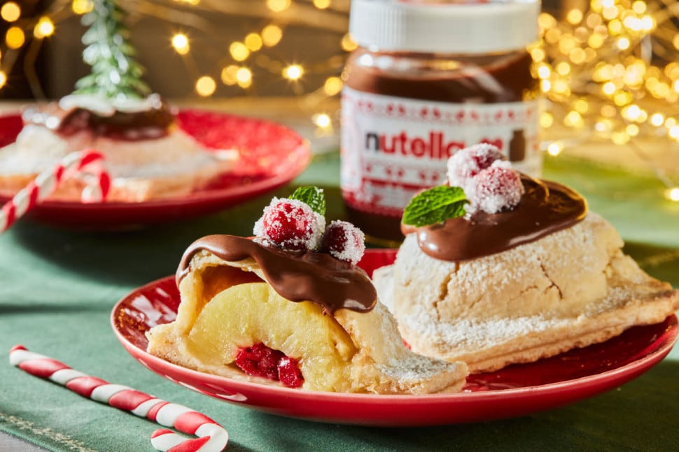 Koláč z rozpůlených jablek na červeném talíři, Nutella® a vánoční výzdoba v pozadí.